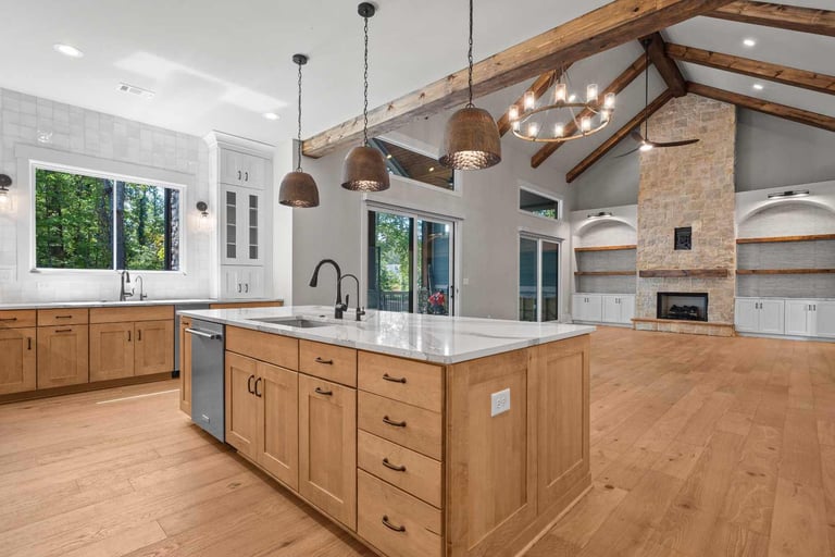 A modern kitchen with wooden cabinetry, a large island with white countertops, pendant lighting, and a view into a spacious living area featuring a stone fireplace and exposed wooden beams. | Paxis Group